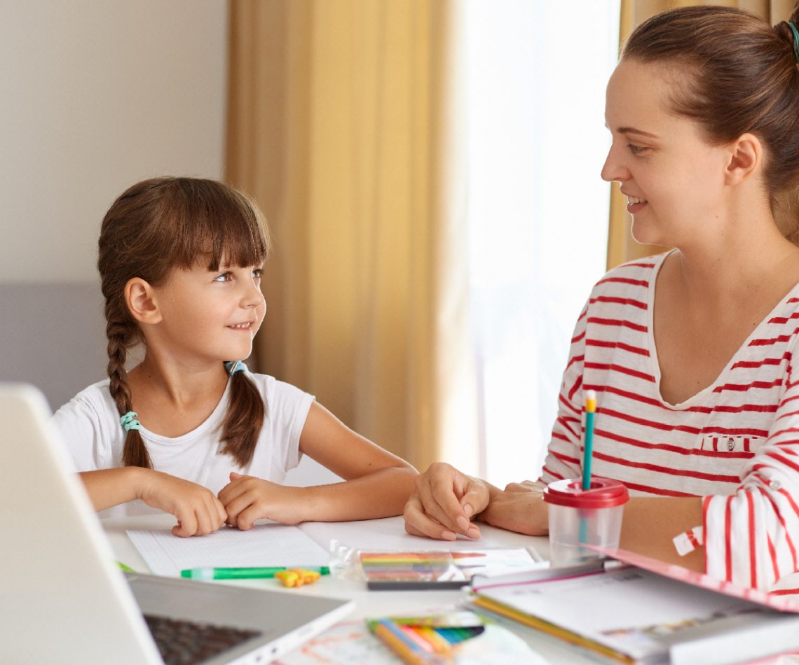 Positive woman with her female kid posing in living room at table, mother helping daughter with lessons, explaining new rule, online distance education.