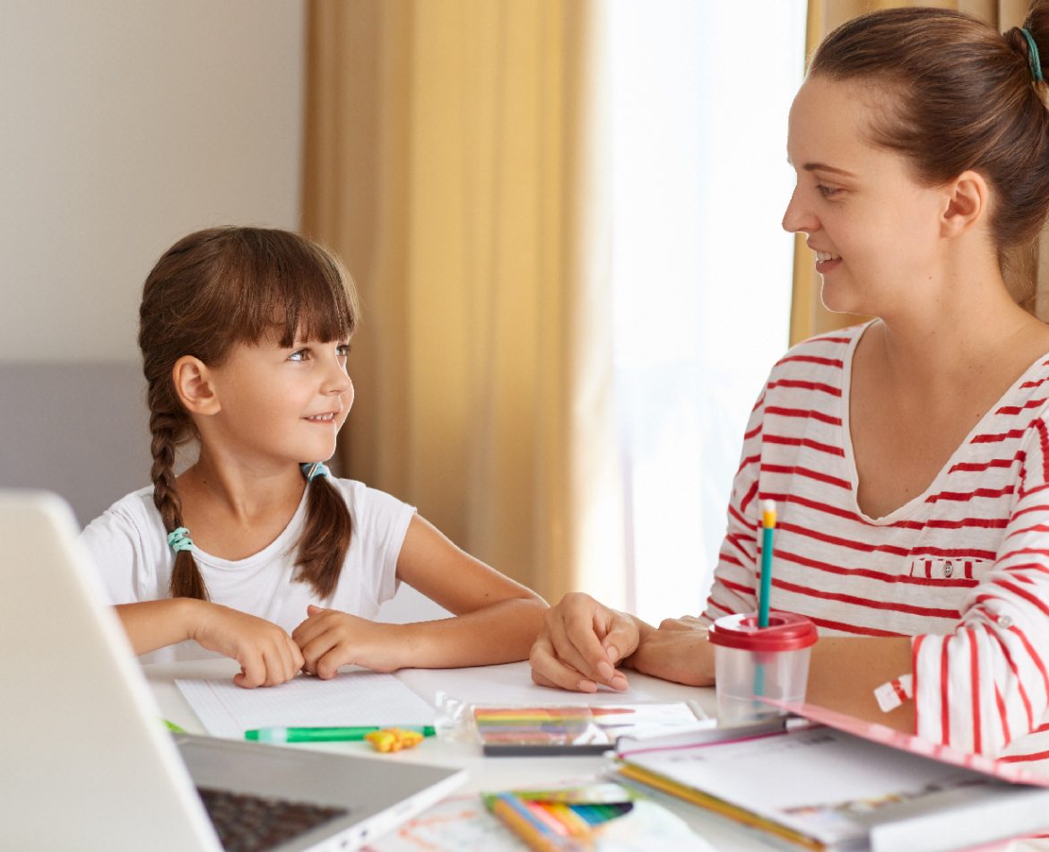 Positive woman with her female kid posing in living room at table, mother helping daughter with lessons, explaining new rule, online distance education.